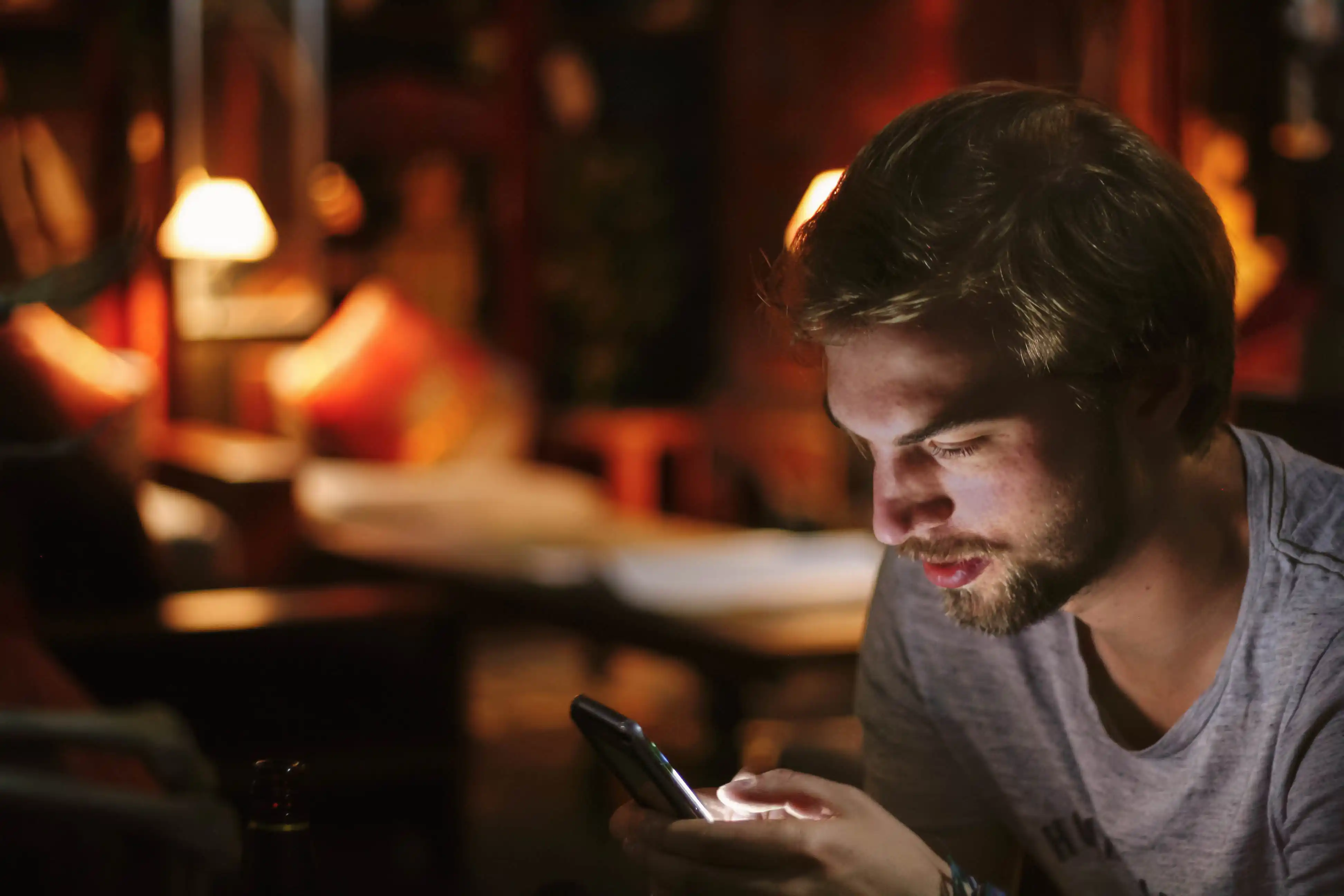 Ein junger Mann mit Bart, der in einem schwach beleuchteten Raum auf ein Smartphone schaut, das Licht des Bildschirms beleuchtet sein Gesicht.