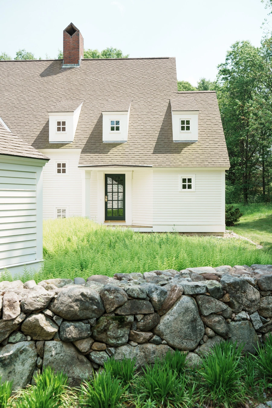 Traditional New England-style home designed by Benjamin Nutter Architects, with stone wall and lush greenery