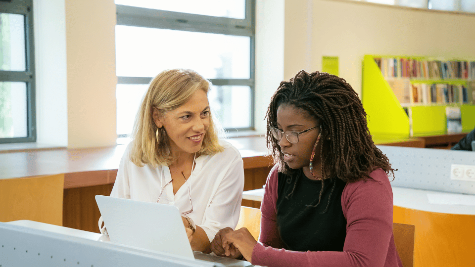 Two women at a library table, focused on a laptop, exemplifying mentorship and professional development
