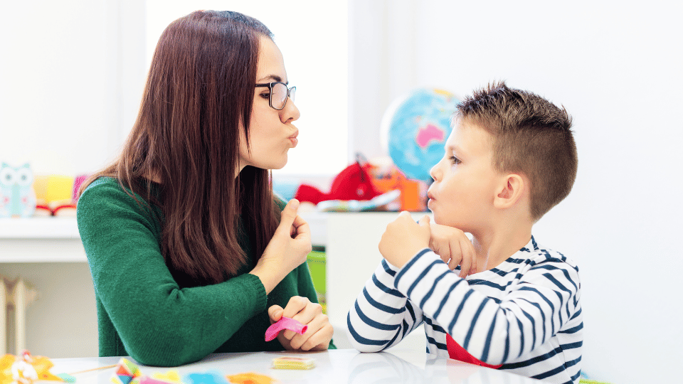 A woman and a boy sit at a table, playing with toys, illustrating a pediatric speech therapy session