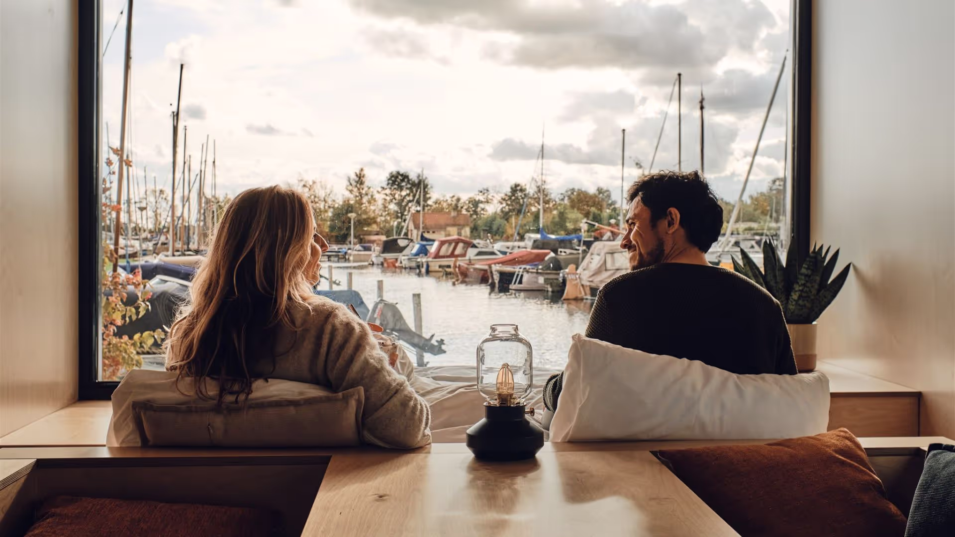 Twee personen zitten in bed voor het grote raam van een Havenhuisje.
