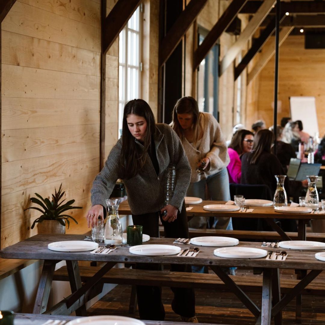 Vrouwen die een lange tafel dekken in een houten zaal