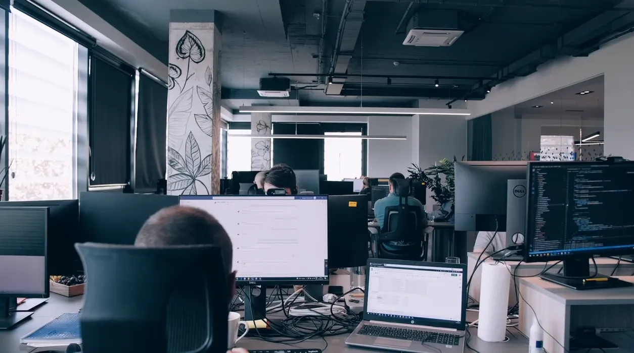Modern office workspace with multiple employees seated at desks, working on desktop computer monitors and a laptop, surrounded by office equipment and some decorative plant and wall art.