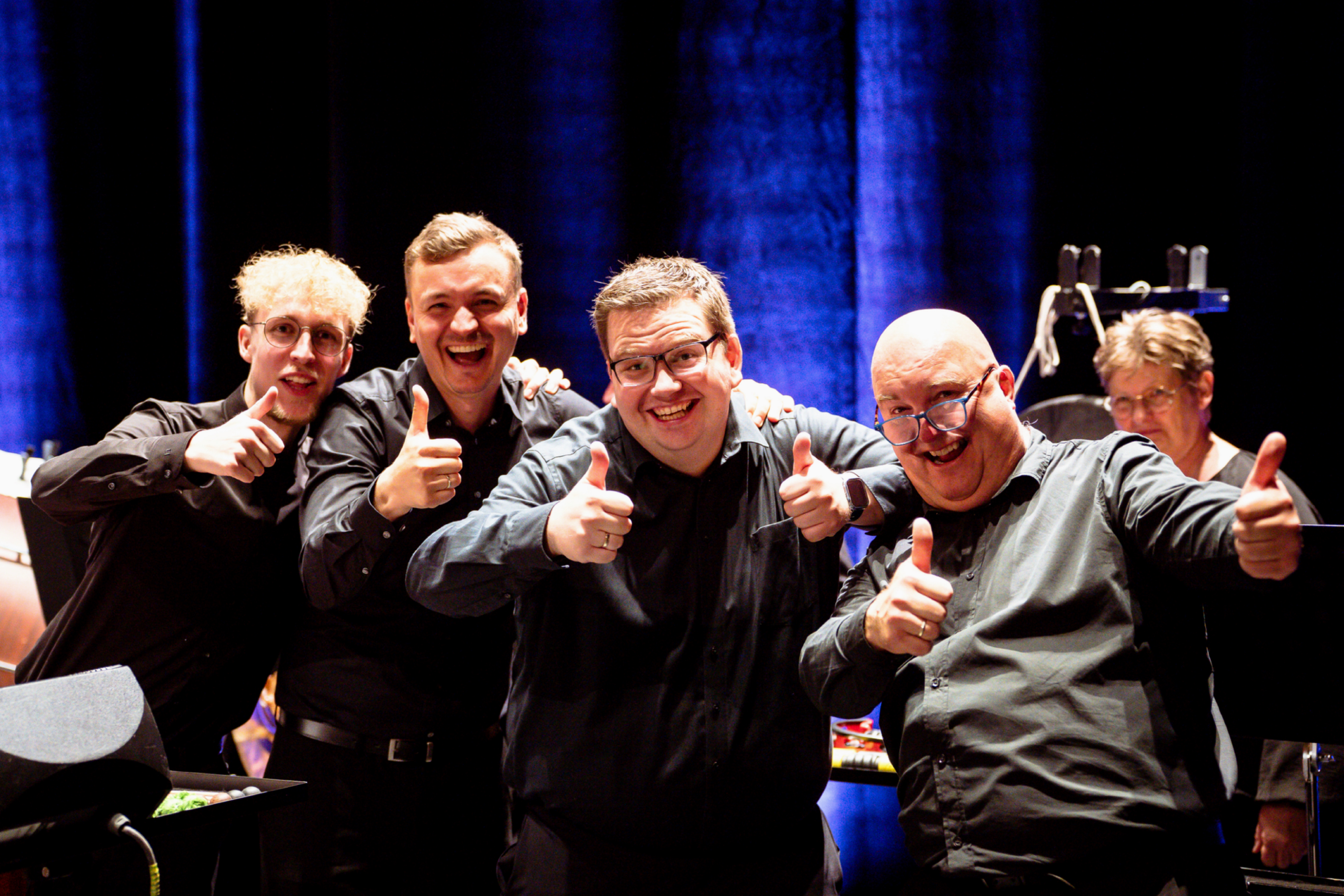 Mehrere Flötistinnen spielen konzentriert während einer Orchesterprobe. Sie sitzen in einer Reihe mit Notenständern vor sich, im Hintergrund sind weitere Musiker zu sehen.