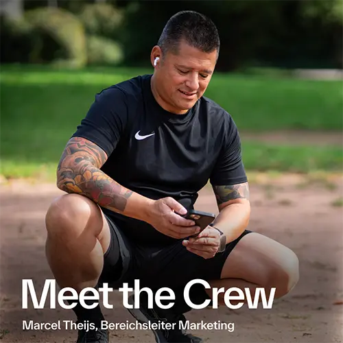 Ein sportlich gekleideter Mann sitzt auf einem Sportplatz und lächelt, während er auf sein Smartphone schaut. Er trägt kabellose Kopfhörer und hat tätowierte Arme. Unten im Bild steht: 'Meet the Crew – Marcel Theijs, Bereichsleiter Marketing'