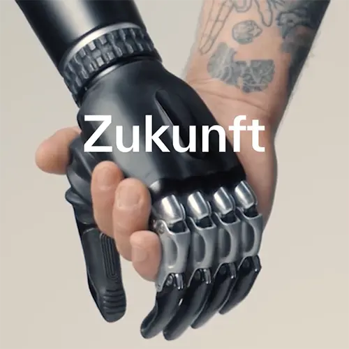 Eine tätowierte menschliche Hand schüttelt eine futuristische, schwarze Roboterhand. Darüber liegt das Wort 'Zukunft' in weißer Schrift.