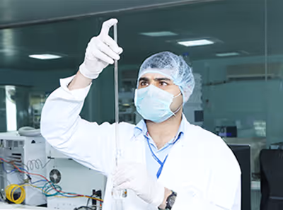 Scientist wearing lab coat, mask, hairnet, and gloves holding a glass pipette in a laboratory.