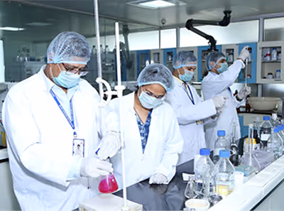 Four scientists wearing white lab coats, hairnets, and masks conducting experiments in a laboratory with various glassware and chemical containers.