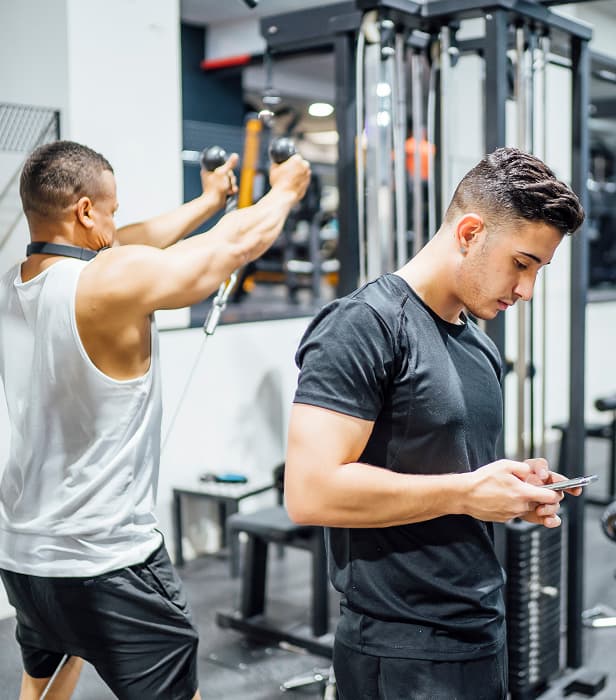Two men in a gym, one exercising with a cable machine while the other checks his phone.