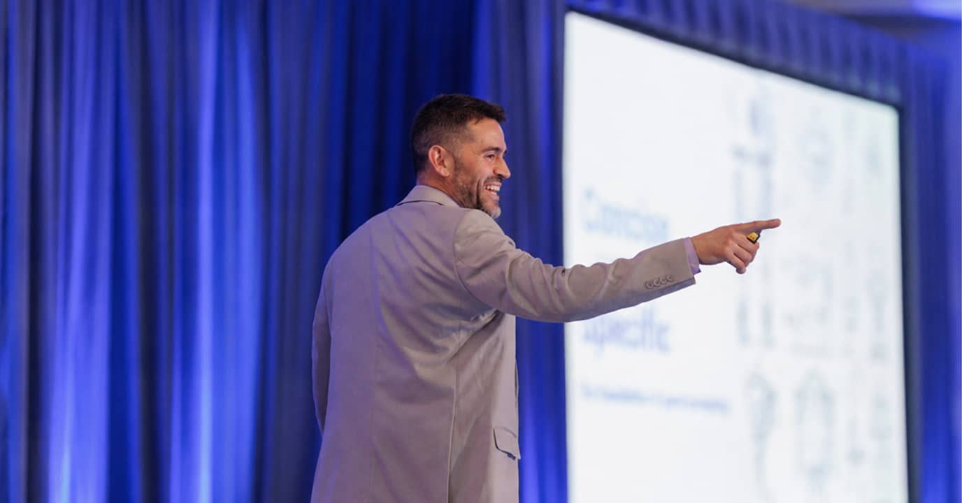 Man in gray suit smiling and pointing while giving a presentation on stage with blue curtains.