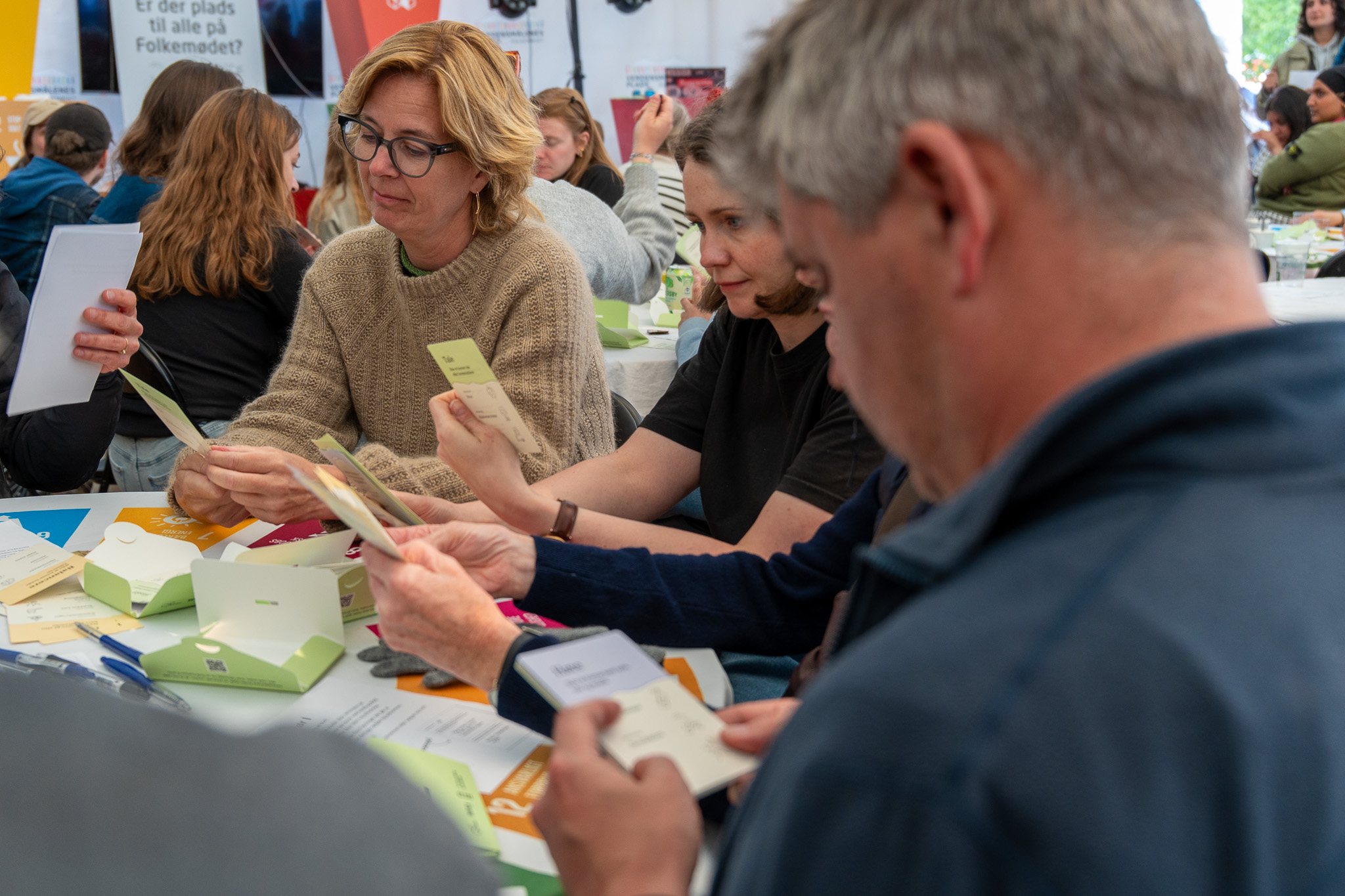 En gruppe mennesker sidder samlet omkring et bord og arbejder koncentreret med kortspilslignende kort og post-its. Kortene har overskrifter som "Universelt Design" og "Funktionsevne" og indgår tydeligvis i en kreativ workshop. Deltagerne bruger pen og papir og peger engageret, hvilket signalerer samarbejde og idéudvikling.