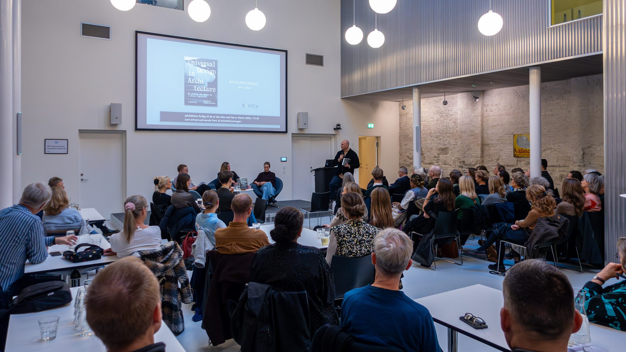 Book launch and panel debate on universal design in a bright auditorium, where a presenter stands at a lectern in front of a large canvas of presentation, while a large audience sits close and listens attentively to the conversation between panelists on stage.