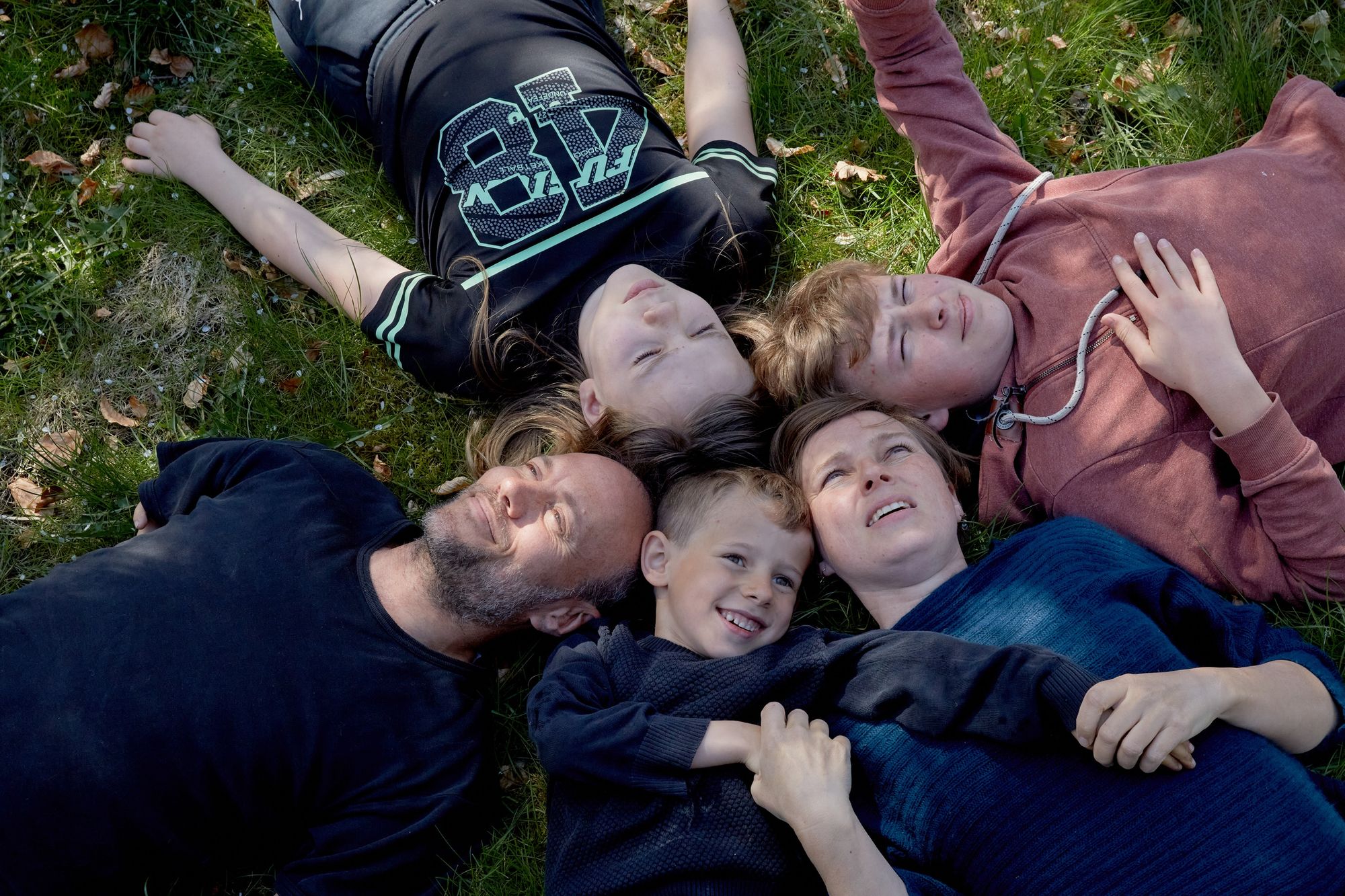 En gruppe mennesker ligger tæt sammen i græsset i en cirkelformation med hovederne mod midten. De ser alle op mod himlen. Lyset falder blødt på deres ansigter, og de ligger afslappet i forskelligt tøj. Stemningen virker varm og tryg.