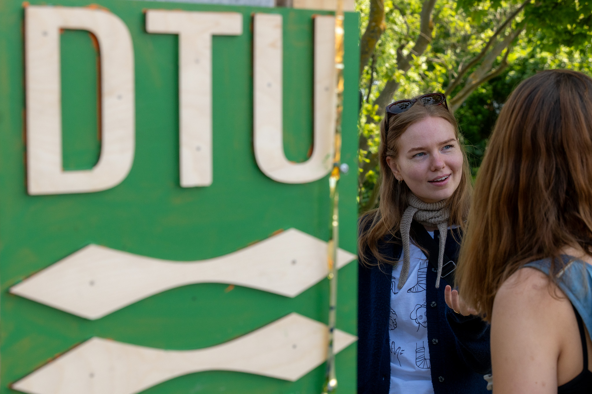 En ung person står ved siden af et stort grønt skilt med bogstaverne ”DTU” og taler med en anden person. Der er træer i baggrunden, og de to ser ud til at være i samtale udendørs.