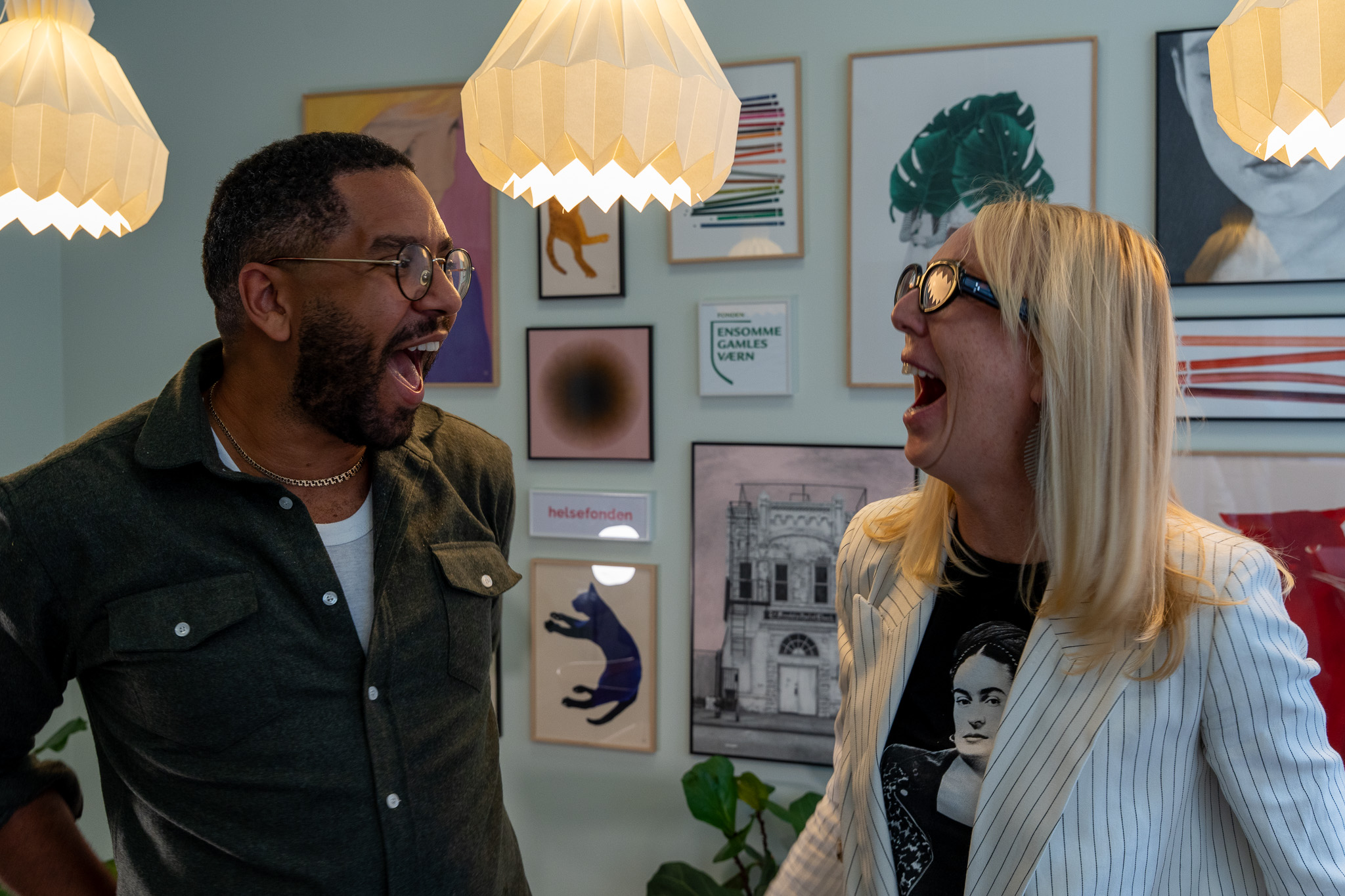 To personer står over for hinanden og griner i et rum med lamper og indrammet kunst på væggen.