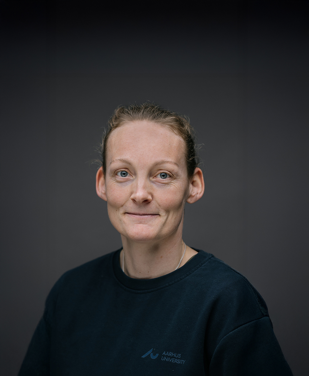Photo of woman looking into the camera with her hair up and a dark blue jersey on where it says Aarhus University
