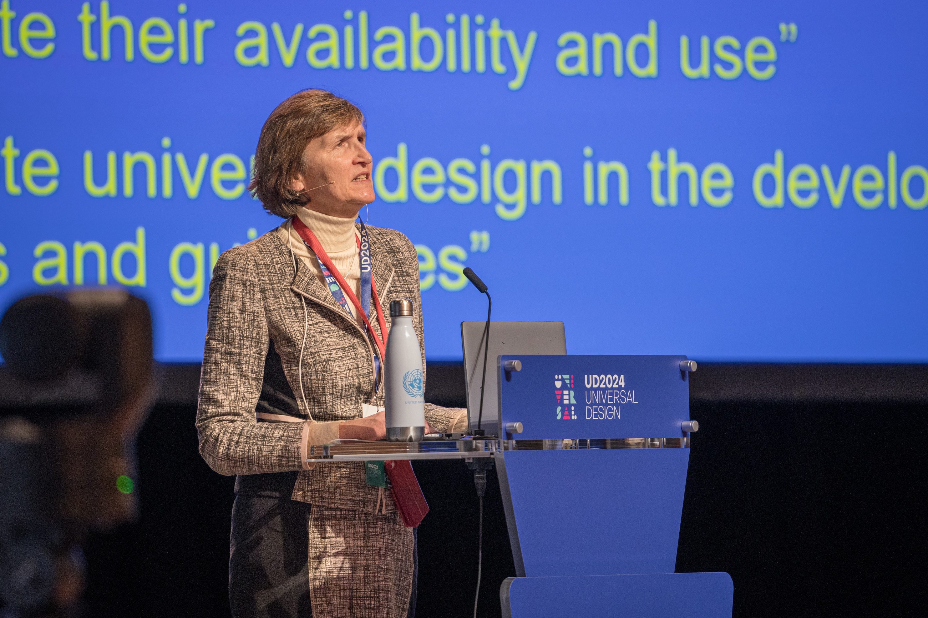 Taler står ved et podie på en konference om universelt design med præsentationsslide i baggrunden og logoet “UD2024 Universal Design” på talerstolen.
