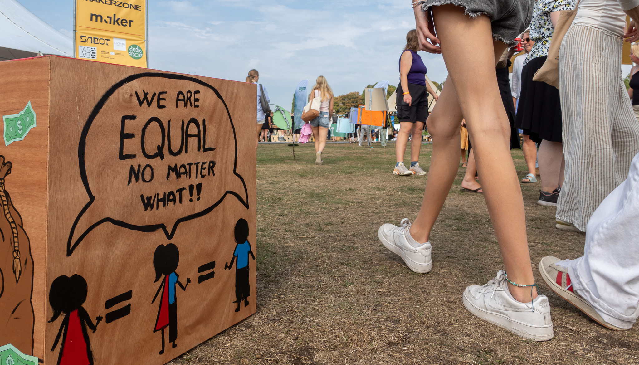 En trækasse med en tegning og teksten “We are equal no matter what!!” står på græsset ved et udendørs arrangement, mens unge mennesker går forbi i baggrunden.