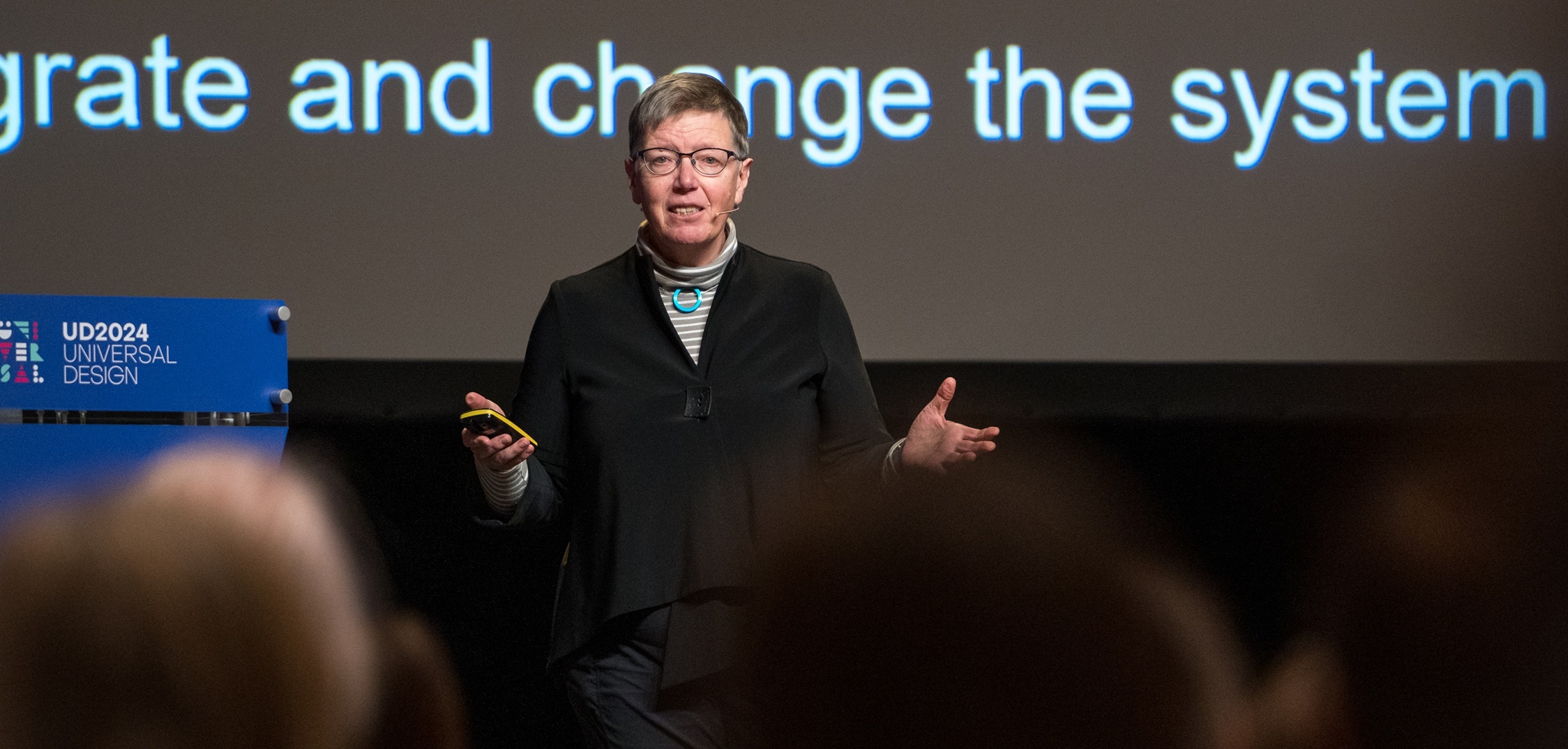 Jutta Treviranus står på scenen og holder oplæg ved UD Oslo 2024 om universelt design, med publikum i forgrunden og en præsentationsskærm i baggrunden.
