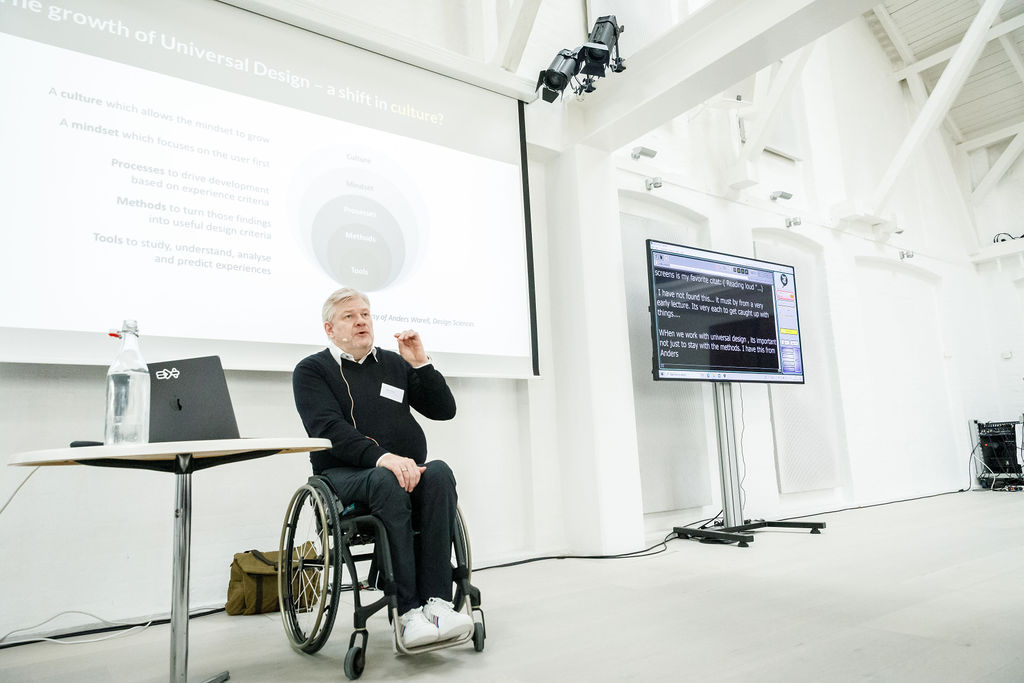 Per-Olof Hedvall siddende i kørestol præsenterer om universelt design i et lyst konferencerum, med slides på storskærm og live-tekstning på en monitor ved siden af.