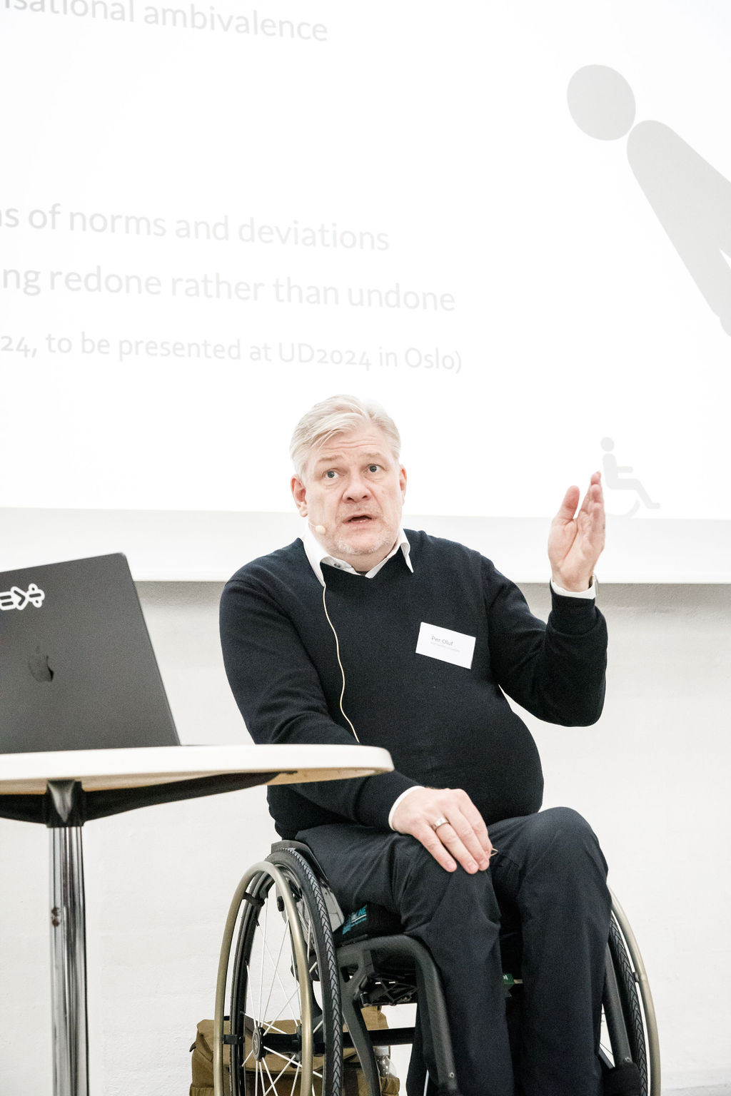 Per-Olof Hedvall siddende i kørestol præsenterer om universelt design i et lyst konferencerum, med slides på storskærm og live-tekstning på en monitor ved siden af.