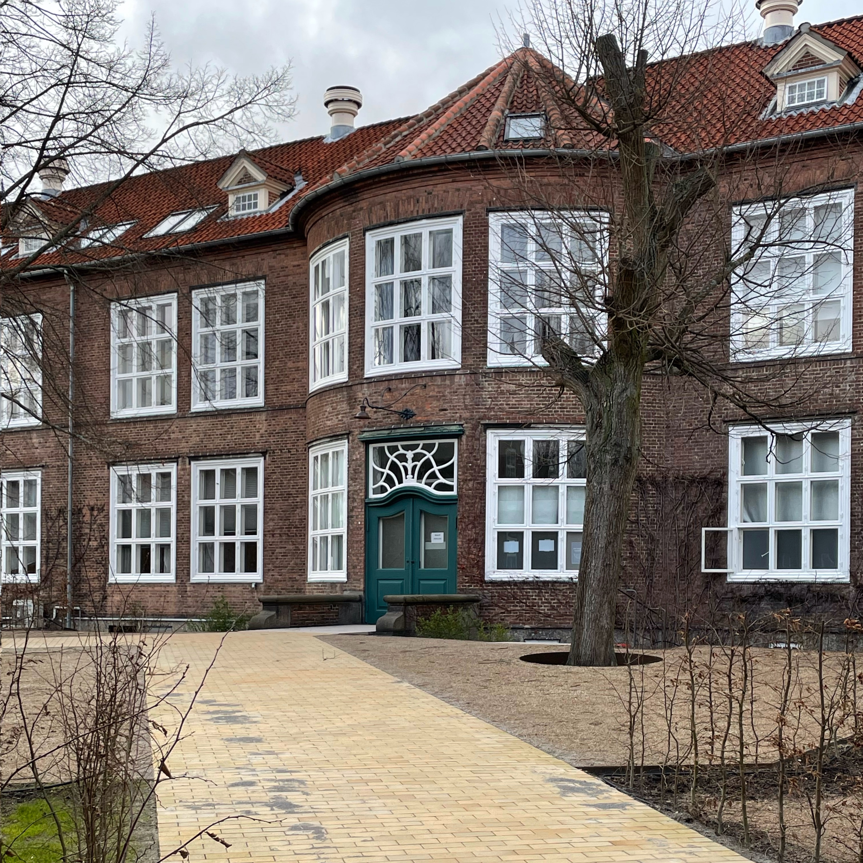 Historisk murstensbygning med buet facade og grøn hoveddør. Foran bygningen fører en bred, jævn sti hen til indgangen.