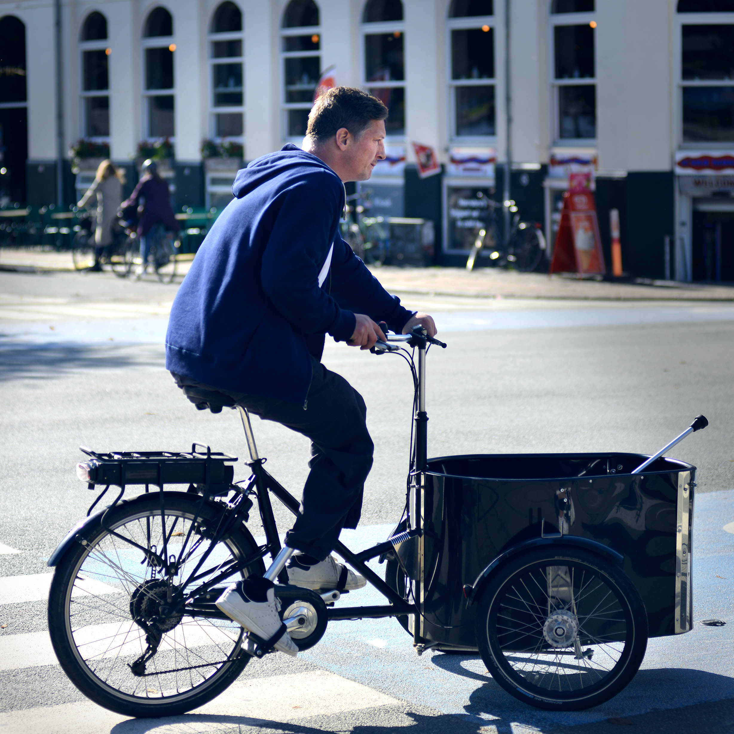 En mand cykler på en sort ladcykel hen over en bygade. Han læner sig let frem, mens han træder i pedalerne, og ladcyklen har en stor ladkasse foran. I baggrunden ses bygninger med høje vinduer og parkerede cykler i dagslys.
