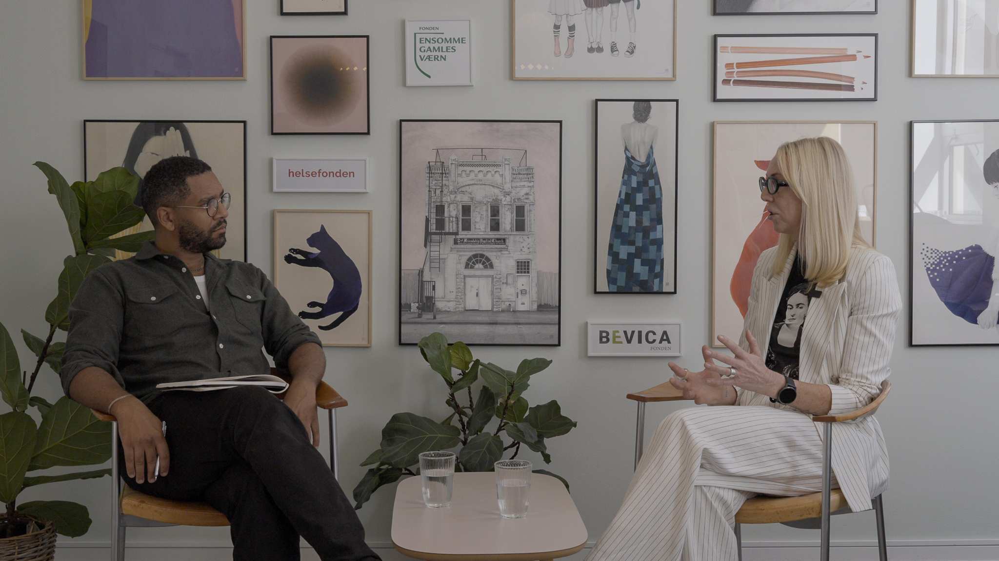 To personer står over for hinanden og griner i et rum med lamper og indrammet kunst på væggen.