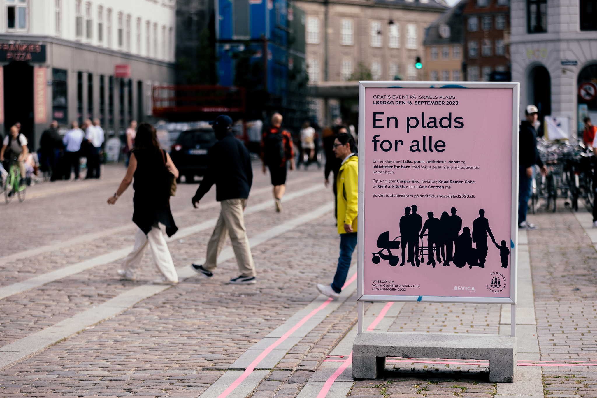 Skilt med teksten “En plads for alle” på Israels Plads i København, mens mennesker bevæger sig gennem byrummet.