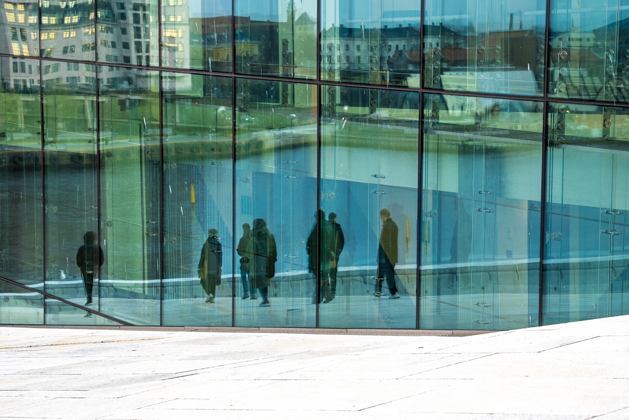 En gruppe mennesker bevæger sig gennem en moderne glasbygning, mens bygningerne udenfor spejles i den store glasfacade.