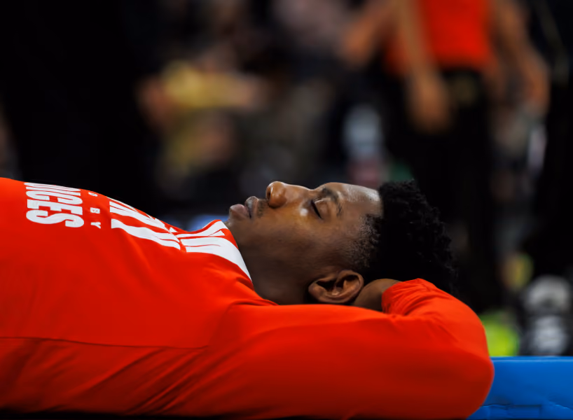RJ Barrett in a red sweatshirt lying down with eyes closed and hands behind head, appearing to rest or relax.