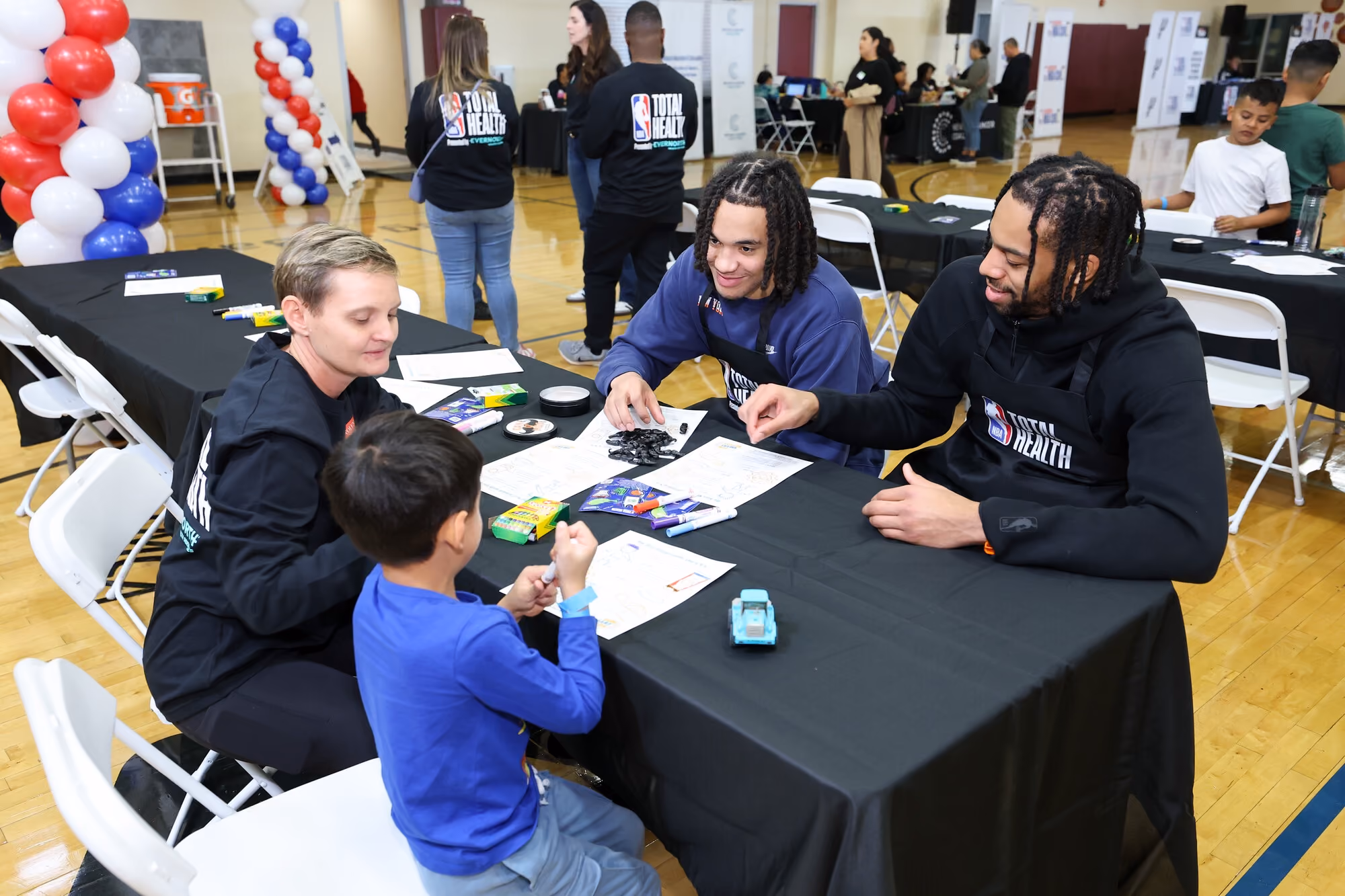 Pacôme Dadiet and Tosan Evbuomwan of NY Knicks at NBA Cup event in a school gym facility with kid and staff.