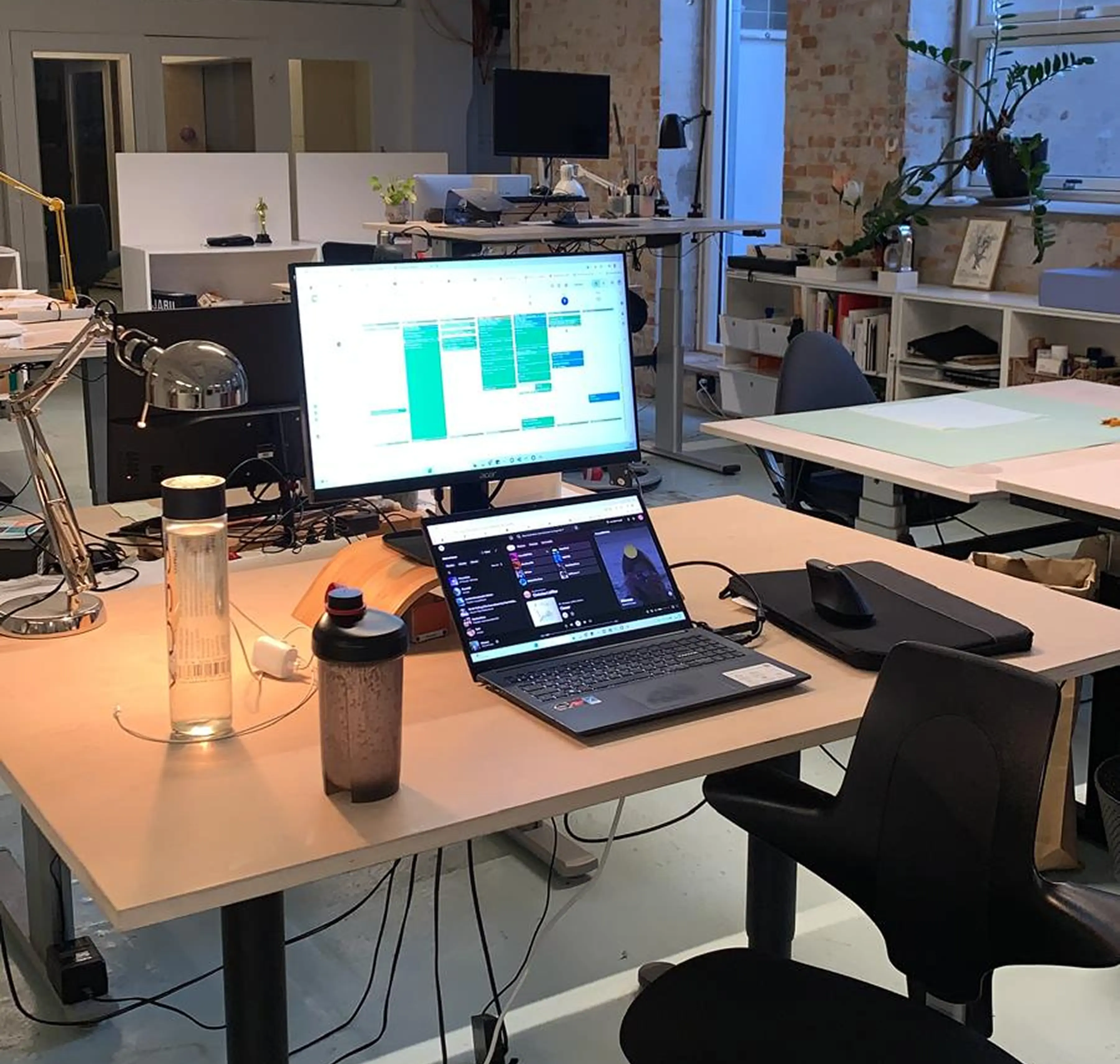 Modern office desk with a laptop, monitor, lamp, water bottle, and office supplies in a bright workspace.