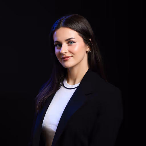 Smiling young woman with long dark hair wearing a black blazer and white top against a black background.