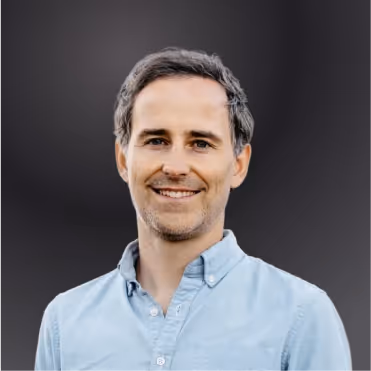 Smiling man with short dark hair wearing a light blue button-up shirt against a dark background.