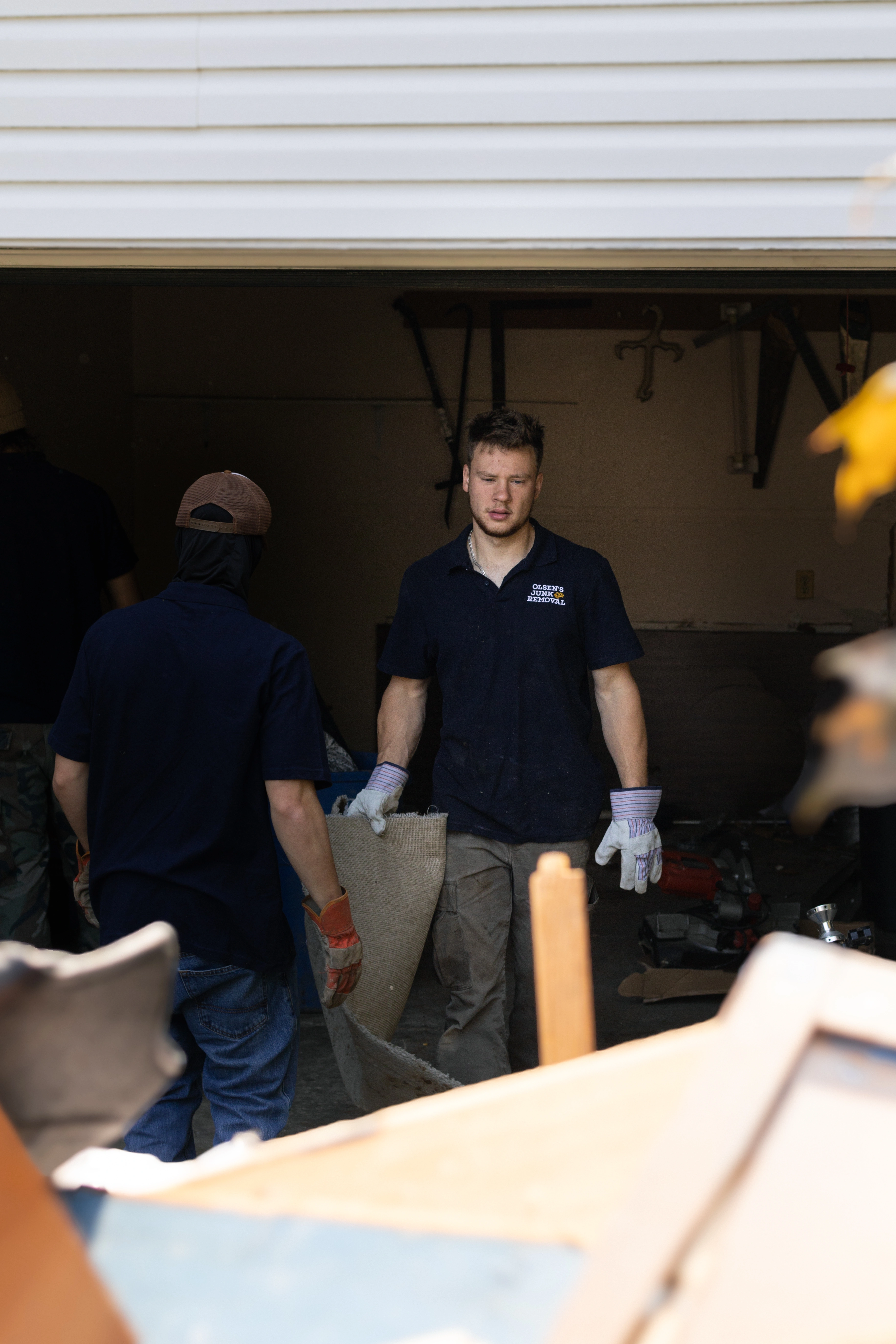 Two men wearing gloves removing a rolled-up carpet from a garage filled with junk.