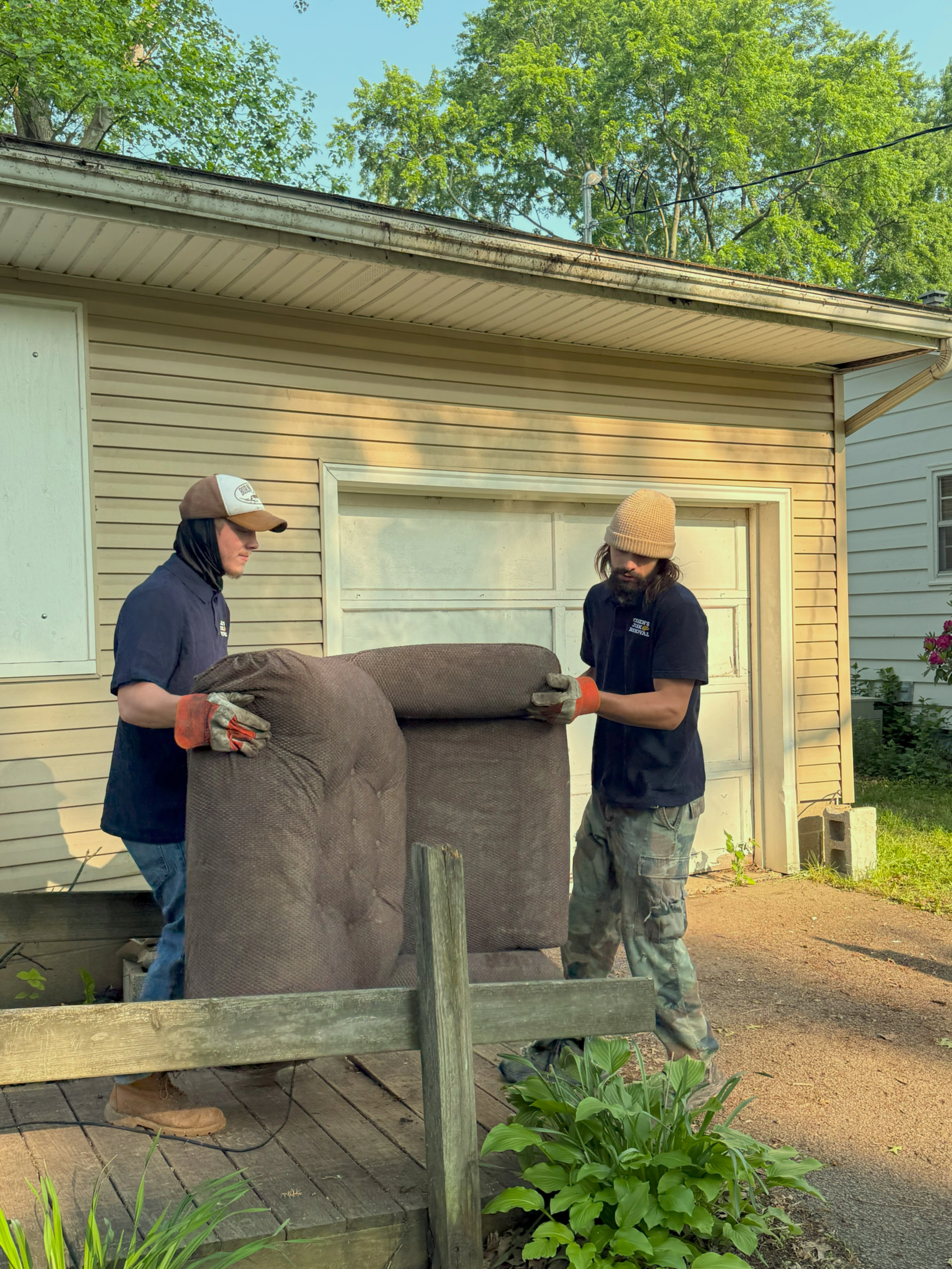 Two men wearing gloves lifting a brown upholstered chair outside a beige garage on a sunny day.