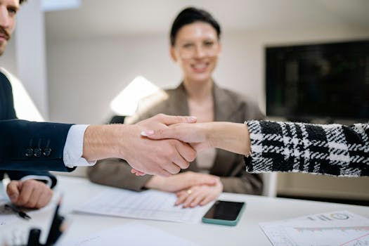 Two individuals shake hands over a desk, symbolizing a successful business agreement. A person in the background looks on, smiling.