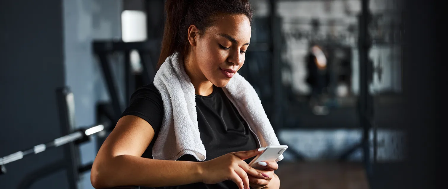 Woman in a gym with a towel around her neck reviewing health data on a digital device