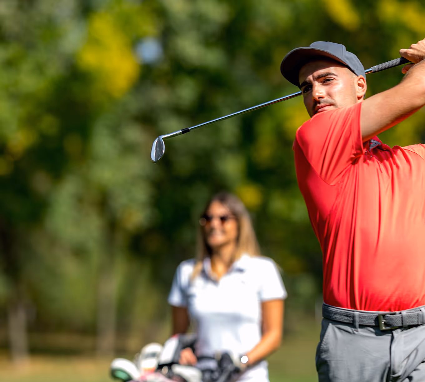 Man in red shirt swinging a golf club with a woman in white standing behind him on a golf course.