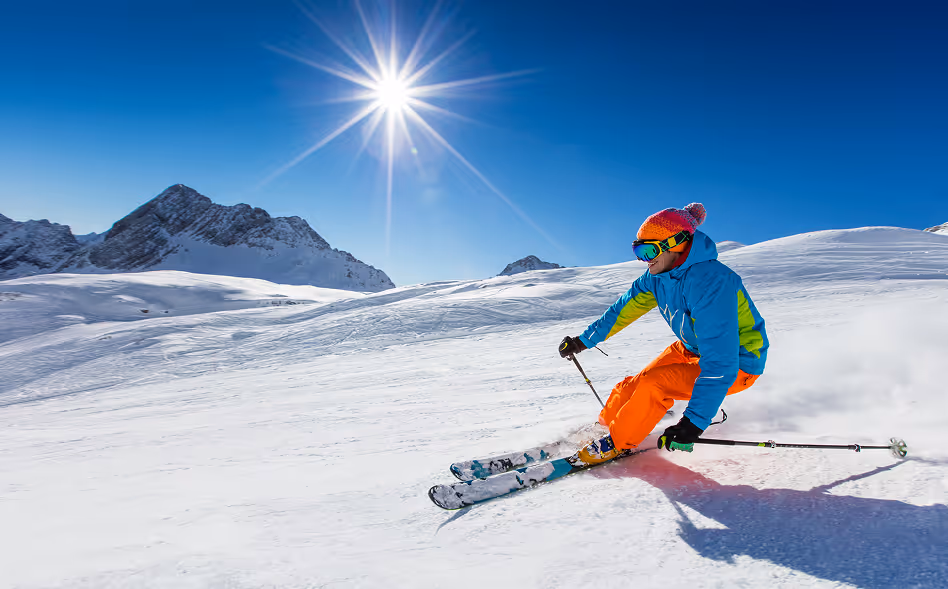 Skier in bright blue and orange gear skiing down a snowy slope under a clear blue sky with the sun shining.