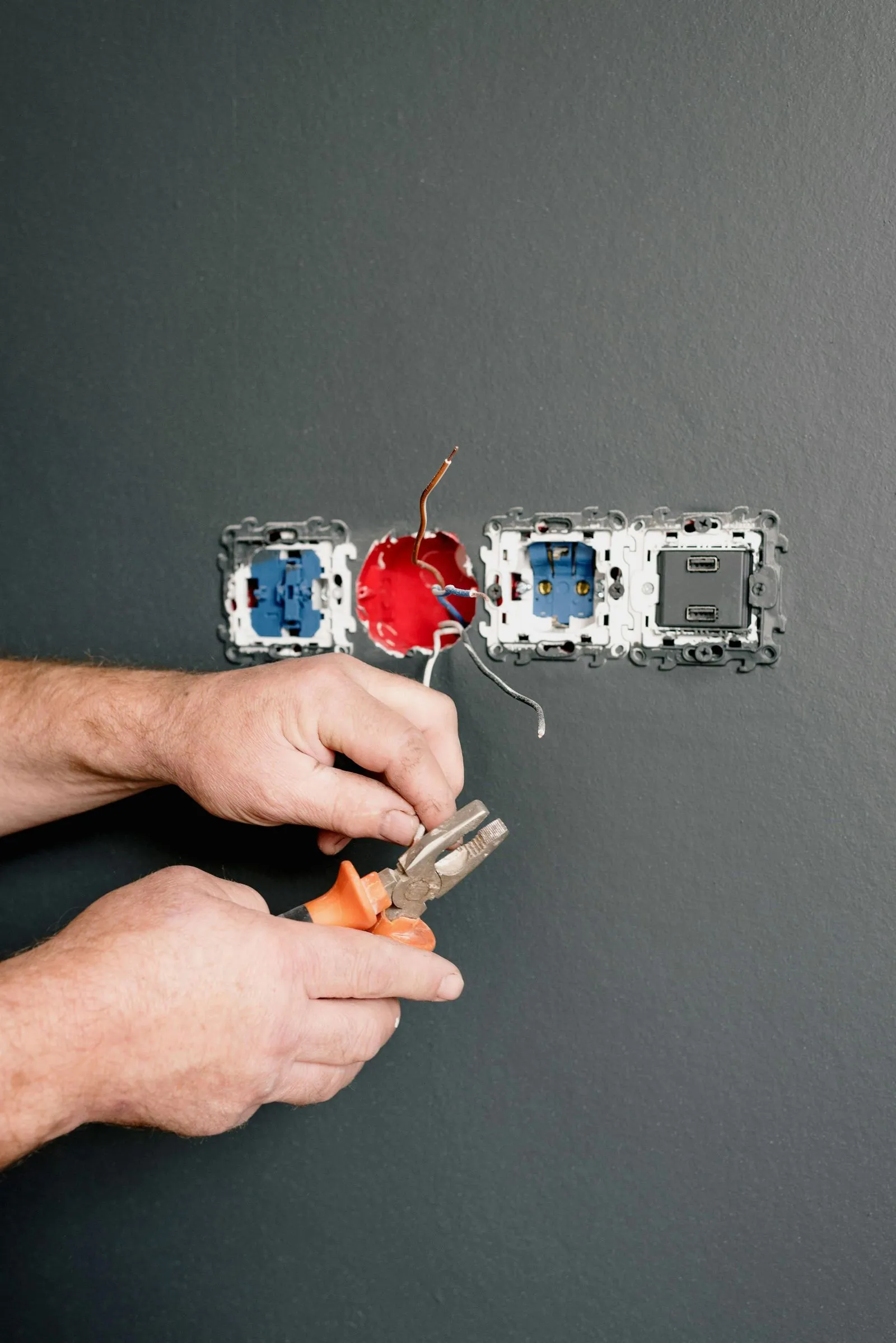 Mains nouant des fils électriques avec une pince devant des prises encastrées dans un mur gris.
