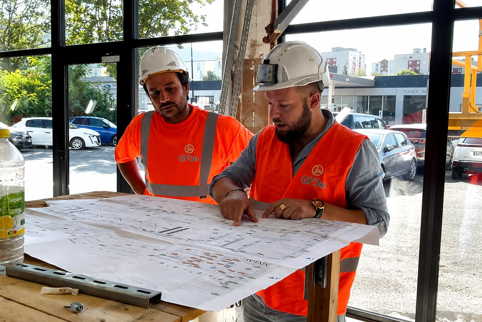 Deux ouvriers en casque et gilet orange examinent des plans sur une table près d'une fenêtre.