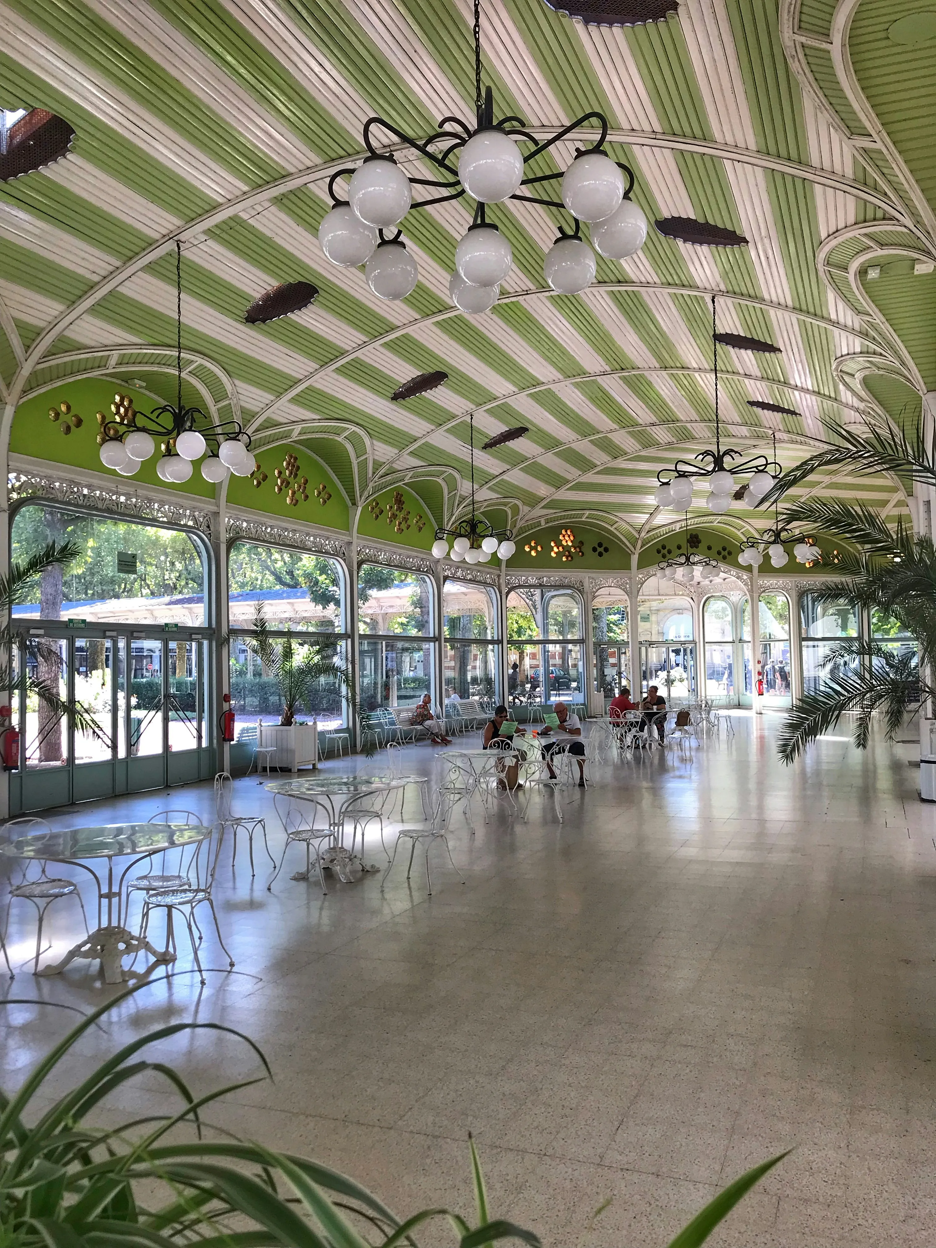 Grand hall with green striped ceiling, ornate chandeliers, glass tables, chairs, and people seated.