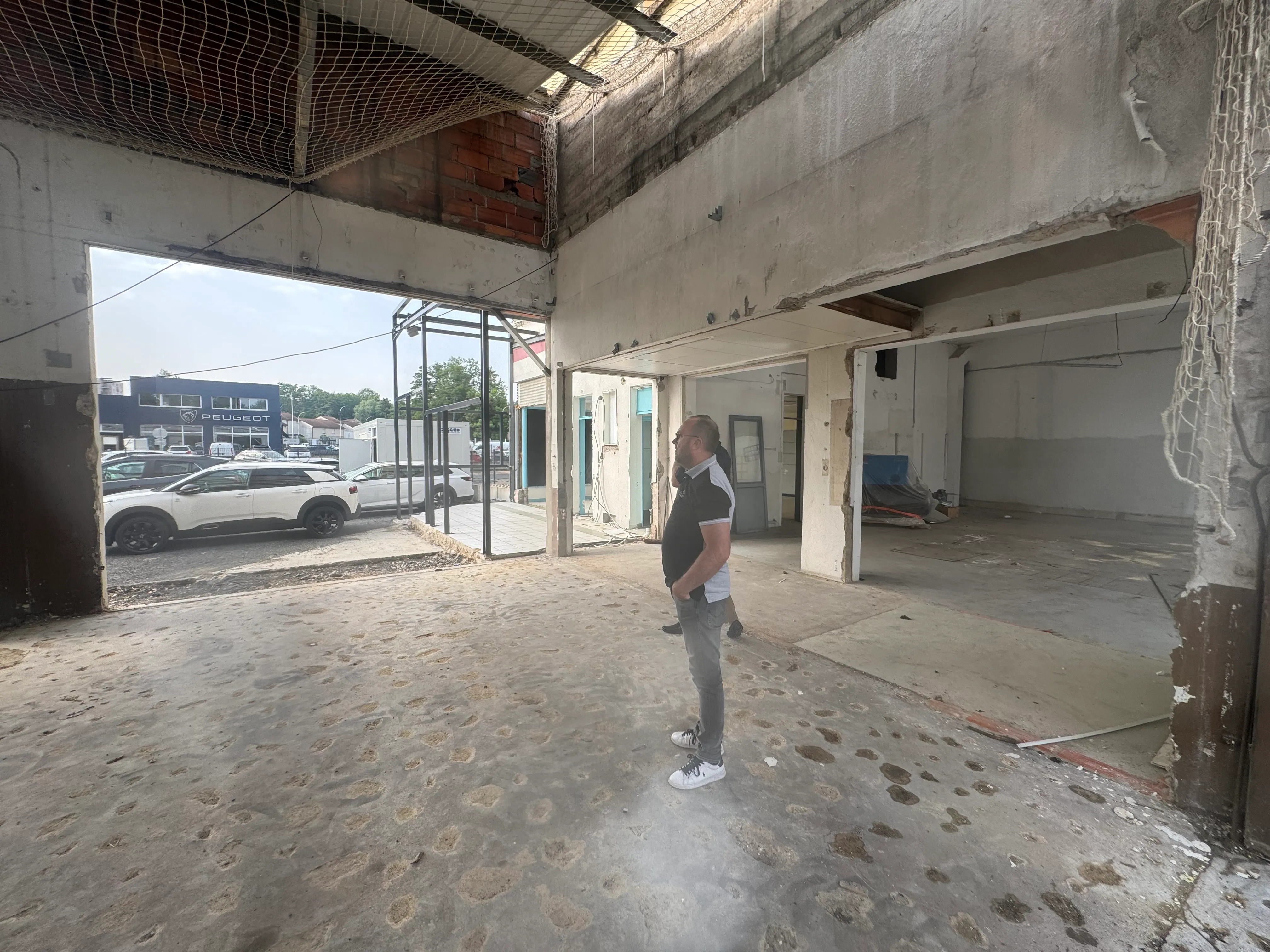 Deux hommes debout dans un espace industriel vide en rénovation, vue sur la rue avec voitures et bâtiment Peugeot.