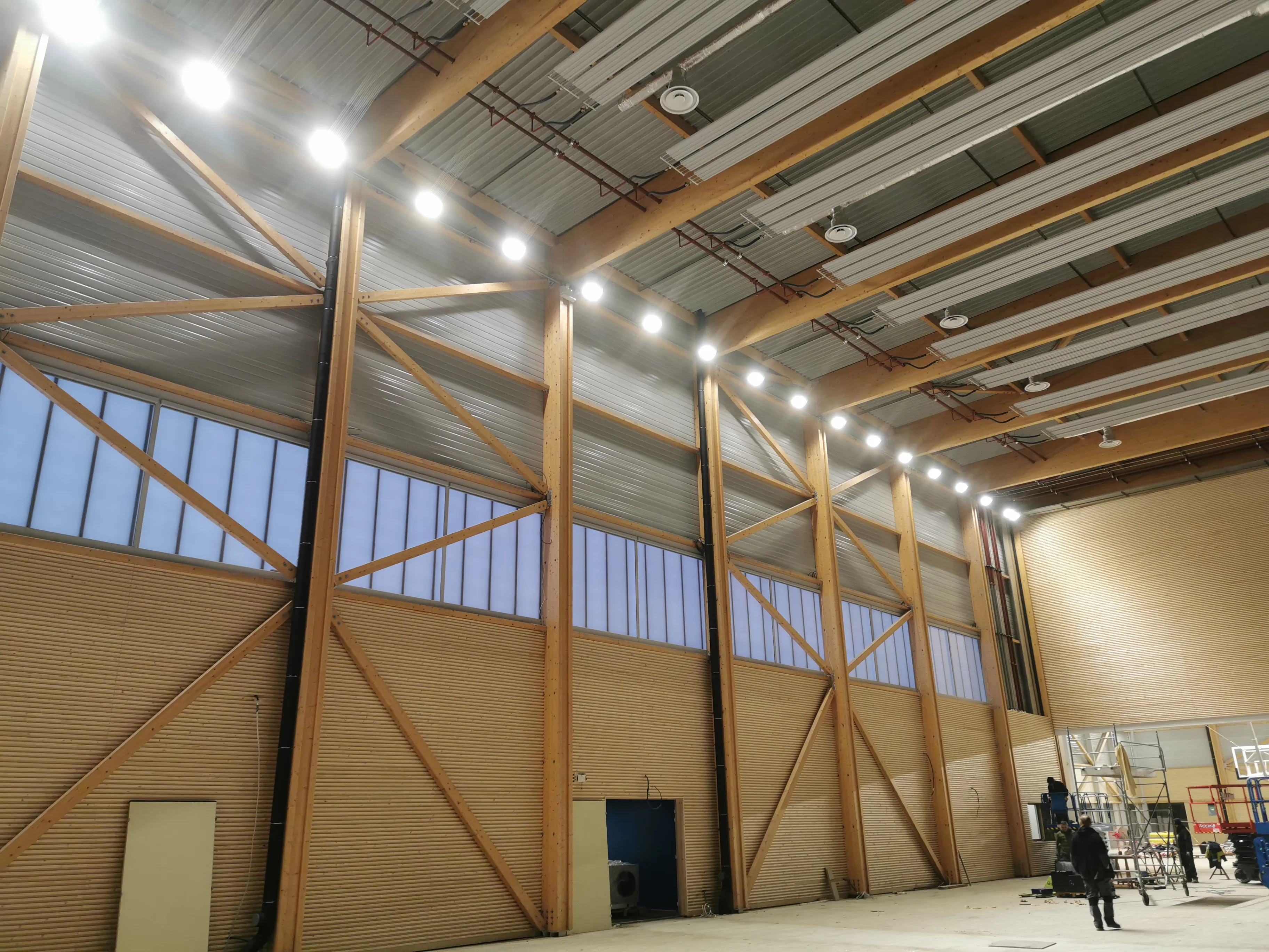 Intérieur spacieux d'un bâtiment industriel avec une grande structure en bois et métal, des fenêtres hautes, un plafond en tôle avec des lumières vives allumées, et des personnes travaillant sur des échafaudages et du matériel au sol.