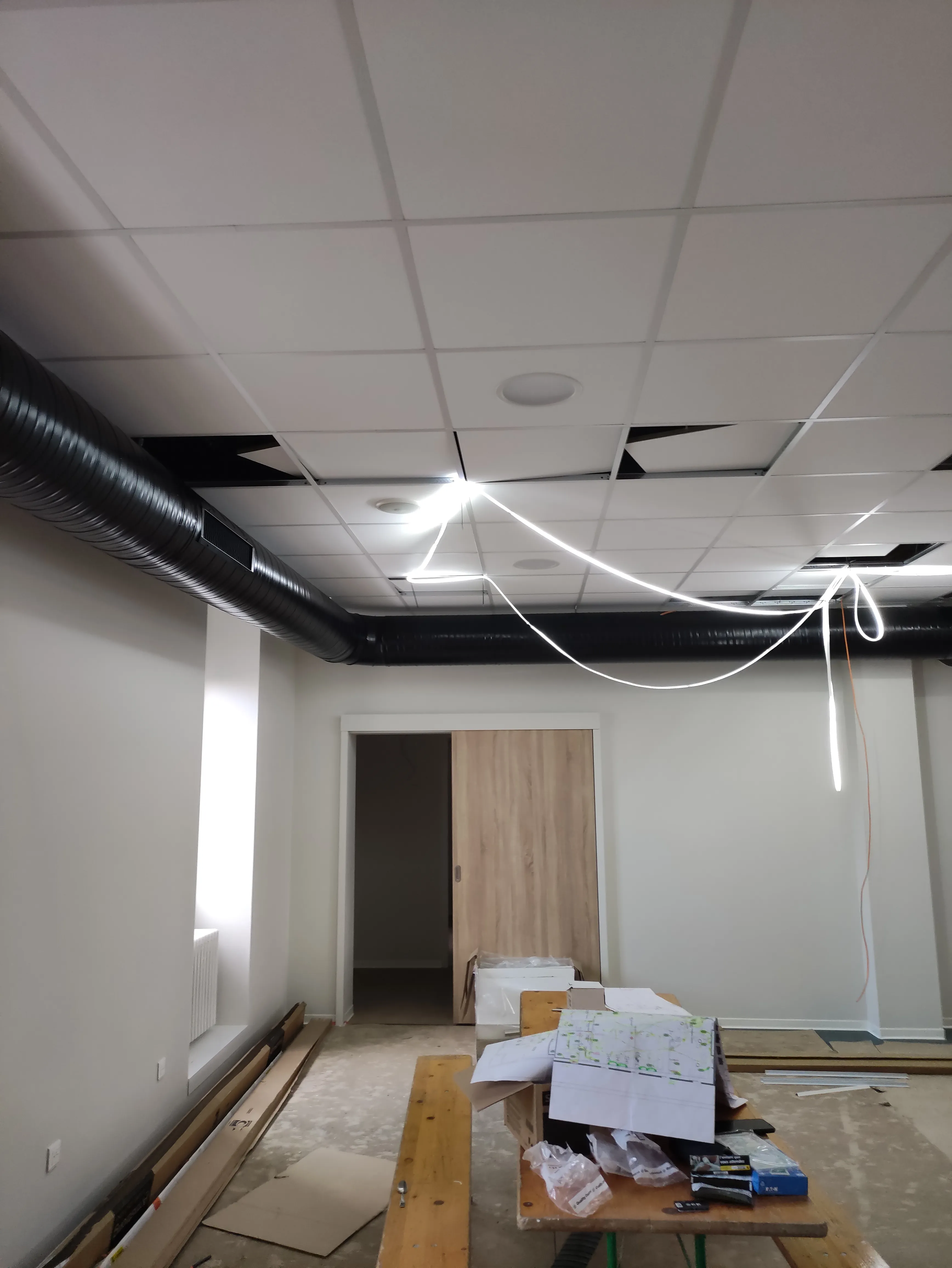Intérieur en rénovation avec des panneaux de plafond suspendu retirés, une grosse gaine noire d'aération, une porte coulissante en bois clair, et une table avec des plans et matériaux de construction posés.