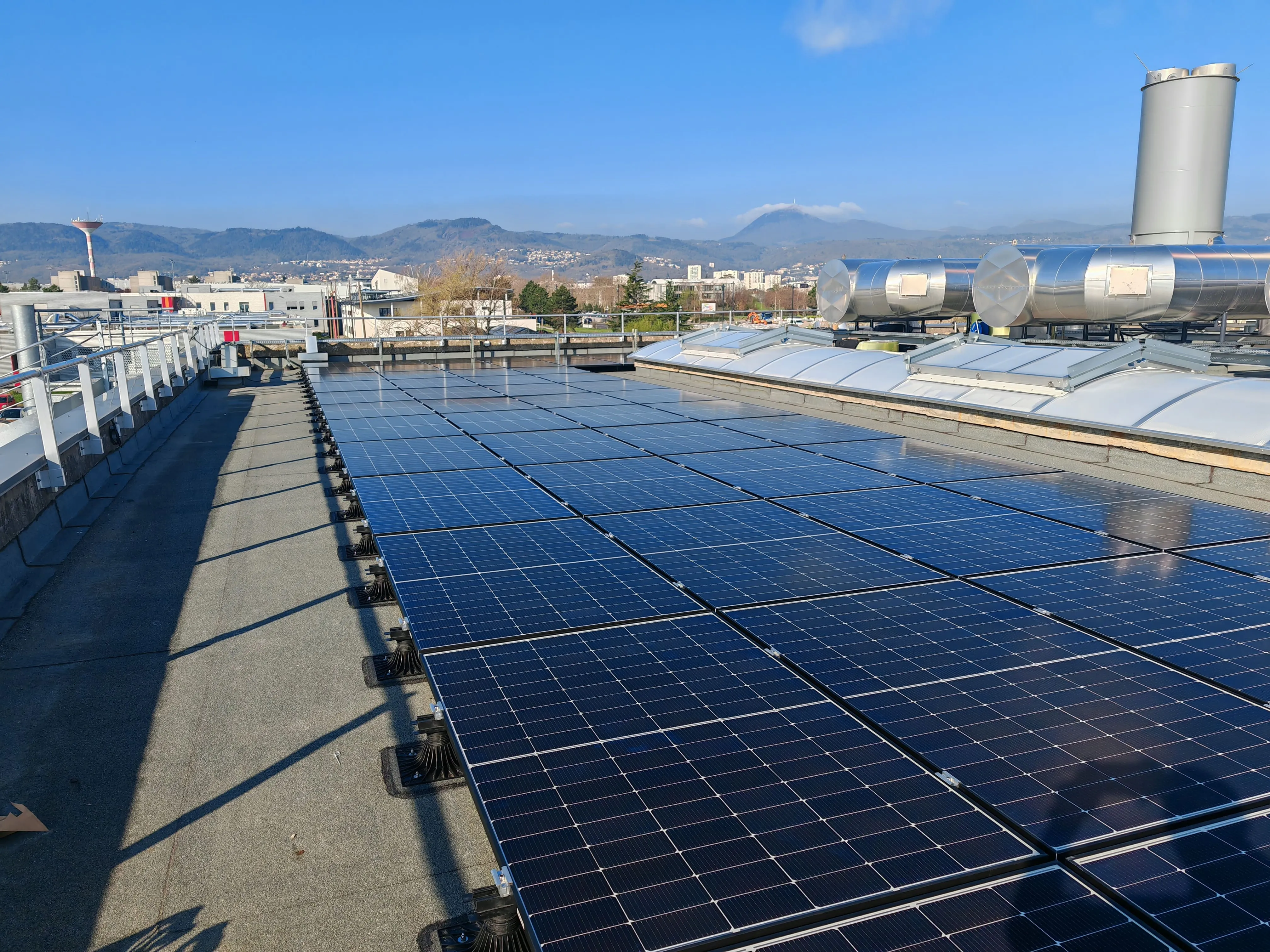 Panneaux solaires installés sur le toit d'un bâtiment industriel avec un ciel bleu clair et une chaîne de montagnes en arrière-plan.