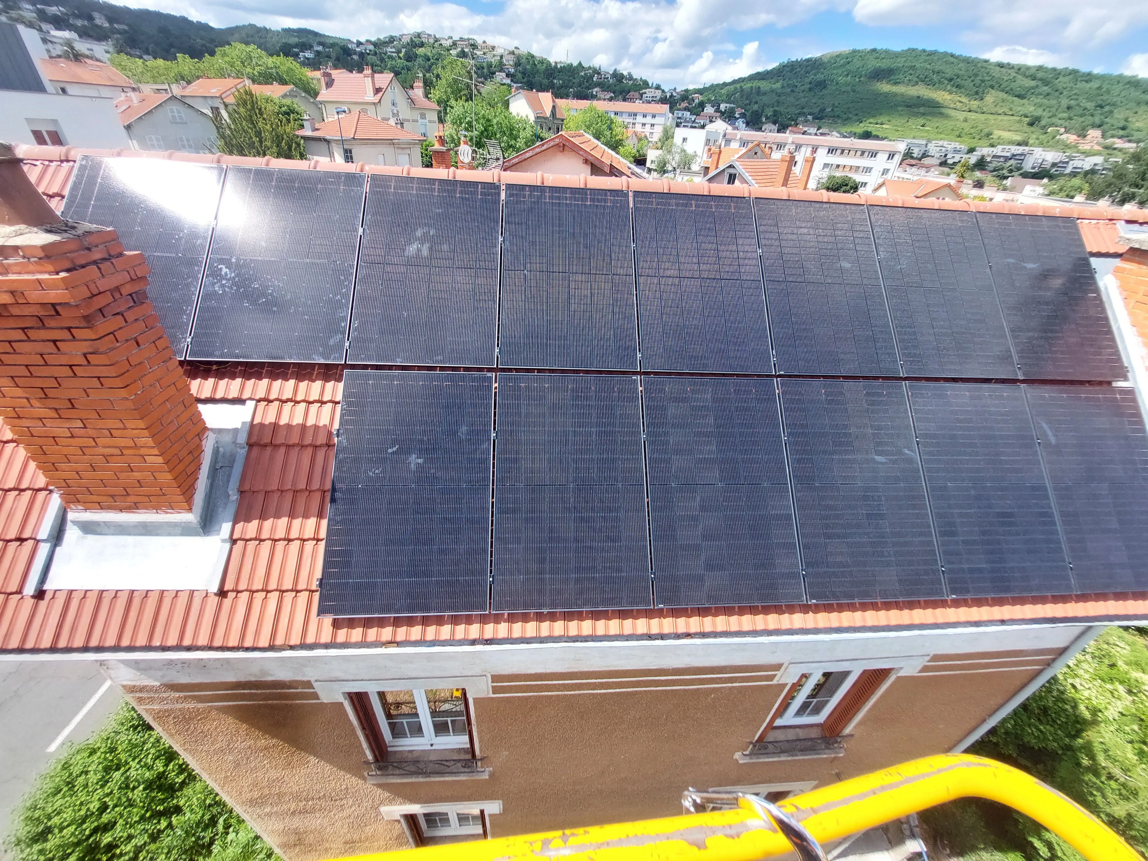 Toit avec des panneaux solaires noirs disposés en deux rangées sur des tuiles rouges, avec une cheminée en brique et une vue de maison et collines végétalisées en arrière-plan sous un ciel partiellement nuageux.