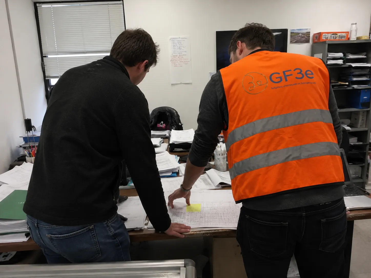 Deux hommes debout, vus de dos, examinent des plans étalés sur une grande table de bureau encombrée de papiers et dossiers, un des hommes porte un gilet orange réfléchissant avec le logo GF3e.